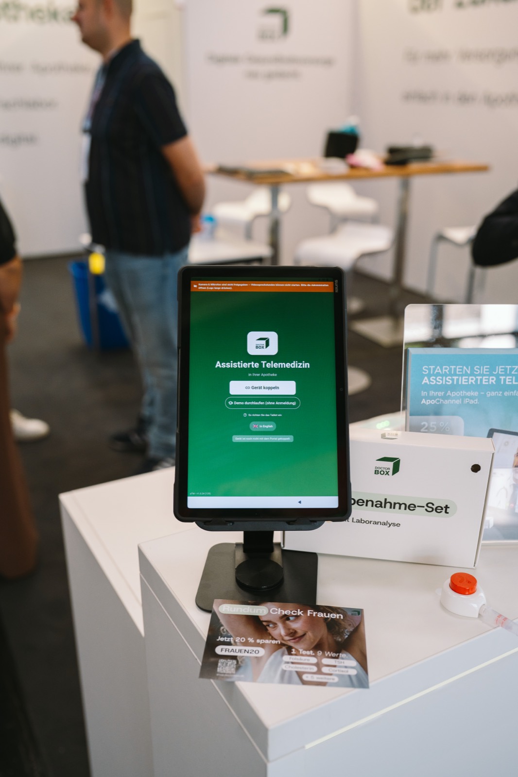 Das Care Tablet auf einer Säule am Stand, auf dem Bildschirm der Startbildschirm der Assistierte-Telemedizin-App